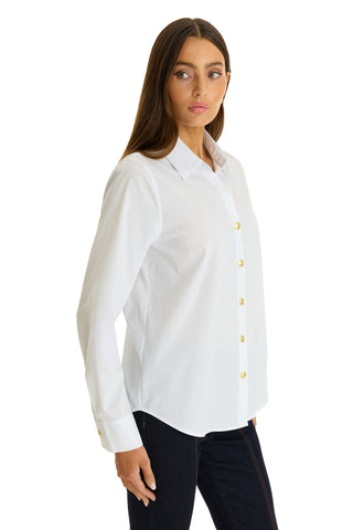 A woman with long brown hair models the Jasmine Shirt - White, featuring yellow buttons and a pleated back yoke, paired with dark pants. She stands against a plain white background, gazing slightly to the side.