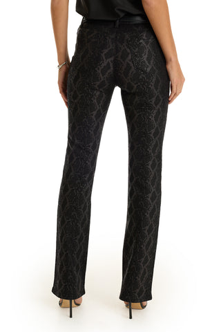 A woman stands facing away, wearing Aria Flare Jean - Black Python pants and black high heels, with her arms relaxed at her sides against a plain white background.