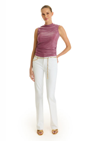 A woman stands against a white background wearing the Mia Shimmer Tank in Amethyst, paired with white pants, a gold chain belt, and beige heeled sandals. Her hair is pulled back and she has one hand in her pocket.