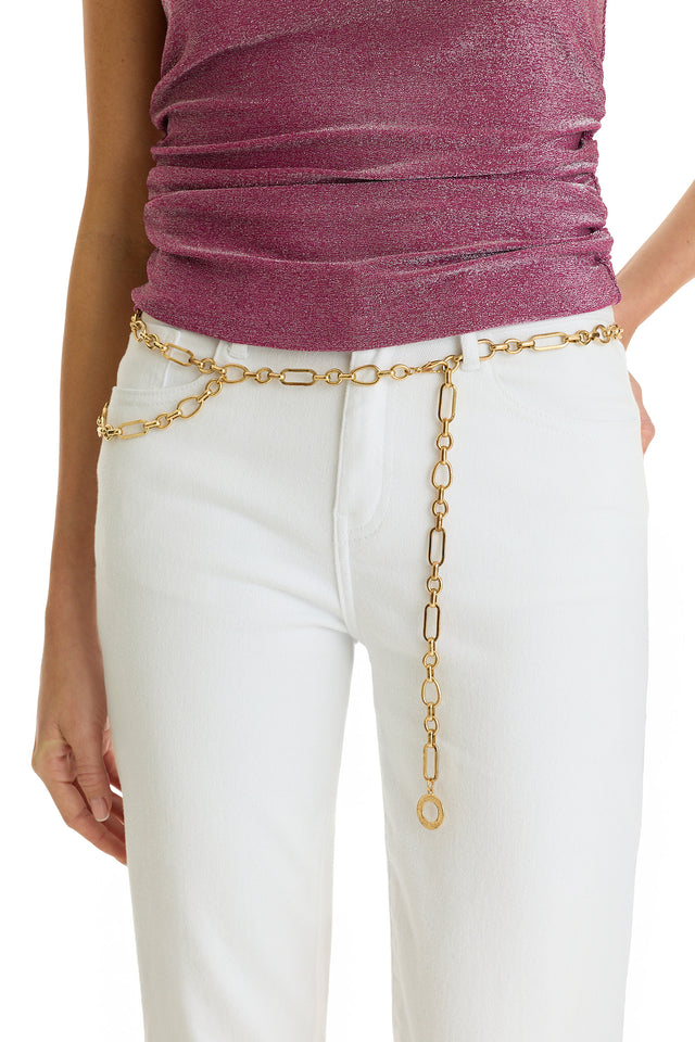 A person wears the Arden Chain Belt - Gold over white pants and a shimmery pink top. The photo centers on the torso and hips, showcasing the elegant gold belt’s dangling link details.