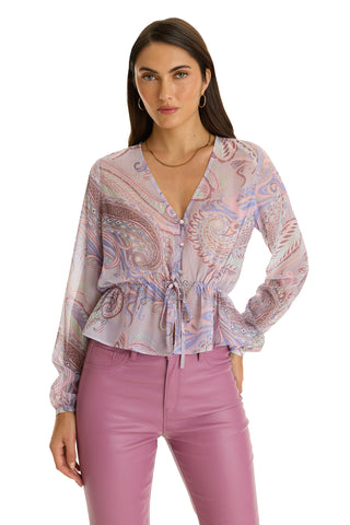 A woman with straight brown hair, facing forward, wears the Whitney Chiffon Top - Pastel Paisley, a V-neck, long-sleeve boho blouse with drawstring waist, paired with high-waisted lavender pants.