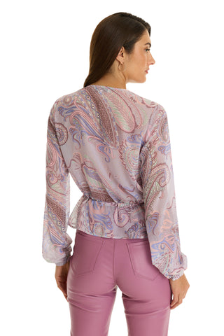 A woman with loose brown hair wears the Whitney Chiffon Top - Pastel Paisley, styled with high-waisted mauve pants and hoop earrings. The sheer, long-sleeved blouse creates a chic boho look as she faces away.