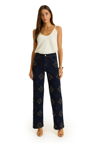A woman faces forward in a white sleeveless top and the Kaia Studded High Rise Jean - Indigo, featuring gold floral stitching. She completes her look with beige heeled sandals, long brown hair, and a neutral expression.