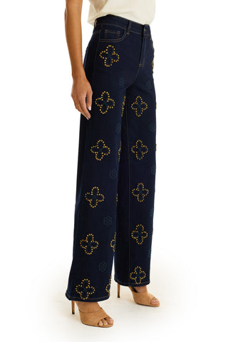 A person wearing the Kaia Studded High Rise Jean in indigo, featuring gold clover studs and a wide-leg fit, styled with a white top and tan heels, stands against a white backdrop.