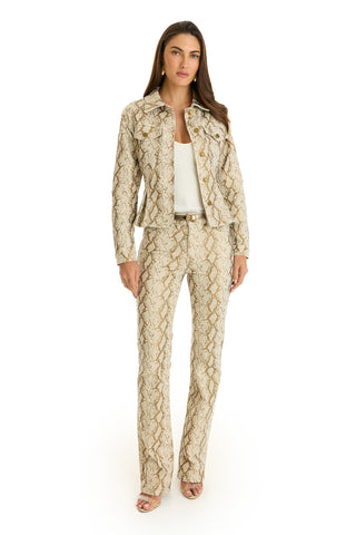 A woman faces forward in the Bethanie Denim Jacket - Gold Python with matching pants, a white top, and open-toed heels. She has long brown hair and a neutral expression, set against a plain white background.