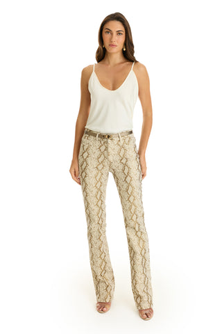 A woman with long brown hair stands facing forward, wearing a white sleeveless top, the Aria Flare Jean - Gold Python with a belt, and open-toed heels. The background is plain white.