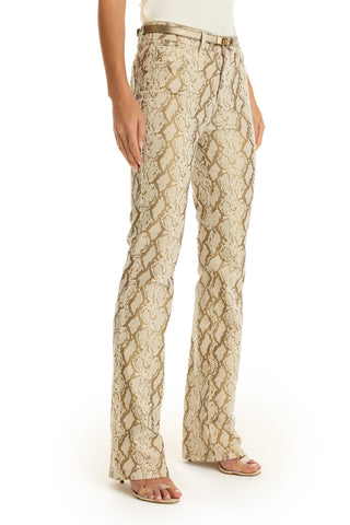 A person models the Aria Flare Jean - Gold Python with a slim bootcut, paired with a white top and beige heeled sandals against a white background. Only the lower half is shown.