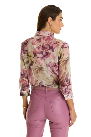 A woman with long brown hair wears the Sara Chiffon Blouse - French Garden, a lightweight floral top in pink, purple, and green tones, paired with high-waisted light purple pants. She stands facing away to show the back of her outfit.