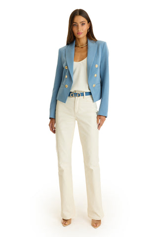 A woman faces forward wearing the Gabrielle Double Breasted Blazer in Serene Blue with gold buttons, a white top, cream pants, beige heels, a gold necklace, and a blue belt. She has long brown hair. White background.