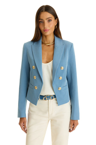 A woman stands against a white background wearing the Gabrielle Double Breasted Blazer in Serene Blue with gold buttons, styled with a white top, cream pants, blue belt, gold hoop earrings, and a gold choker necklace.