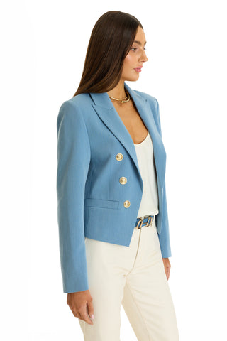 A woman with long brown hair wears the Gabrielle Double Breasted Blazer in Serene Blue, styled with a white top and cream pants. She is shown in profile against a white background.