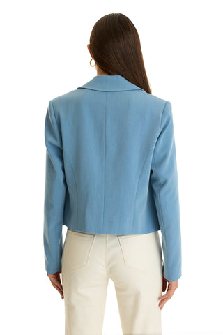 A woman with long brown hair and gold hoop earrings wears the Gabrielle Double Breasted Blazer in Serene Blue and high-waisted cream pants, standing with her back to the camera against a white background.