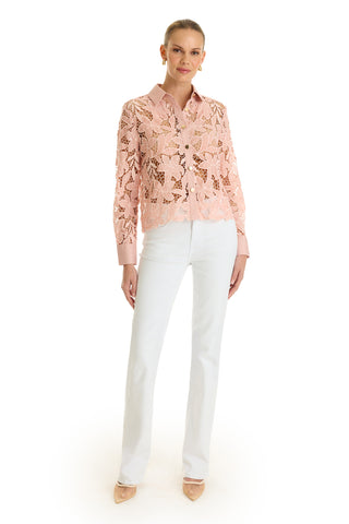 A woman stands against a white background wearing the Mina Lace Blouse in blush over a shirt, paired with white pants and nude heels. Her hair is tied back and she smiles softly at the camera.