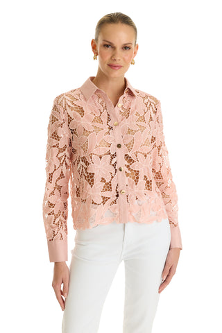 A woman wears the Mina Lace Blouse - Blush, a light pink lace top with delicate floral patterns, long sleeves, and a collar, paired with white pants against a white background.