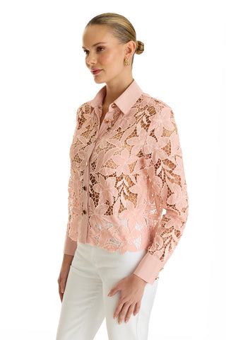A woman stands against a white background wearing the Mina Lace Blouse in blush, featuring delicate floral lace over a matching camisole, paired with white pants. Her hair is in a sleek low bun and she accessorizes with hoop earrings.