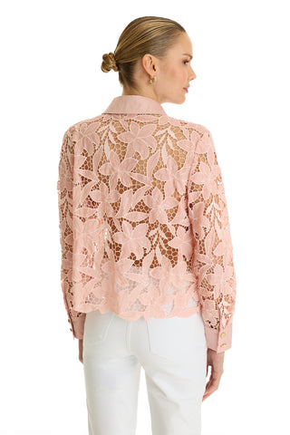 A woman is seen from the back wearing the Mina Lace Blouse in blush—a light pink, long-sleeved lace top with floral patterns and a collar—paired with white pants, set against a plain white background.