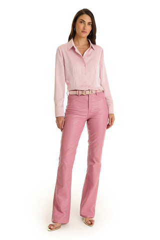 A woman with long brown hair and a neutral expression stands against a white background, wearing the Aria Coated Flare Jean in Paradise Pink paired with a light pink shirt and open-toed heels.