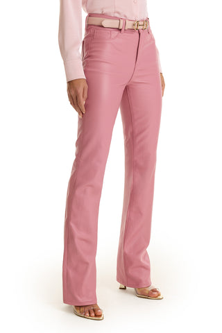 A person stands against a plain white background, wearing high-waisted Aria Coated Flare Jean in Paradise Pink with a matching belt, a light pink button-up shirt, and open-toed heels.