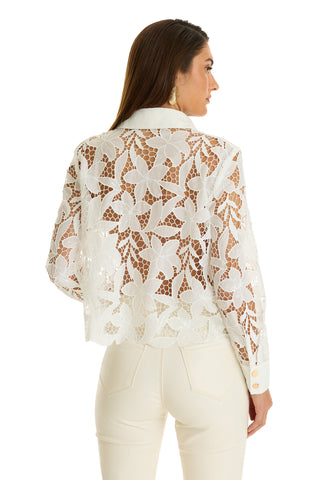 A woman stands facing away, wearing the Mina Lace Blouse - White with long sleeves and high-waisted white pants. Her brown hair flows down, and she accessorizes with gold earrings.