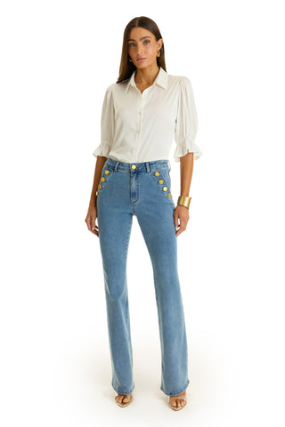 A woman with long straight hair and a neutral expression faces forward, wearing the Cindy Ruffle Sleeve Blouse in white, high-waisted blue jeans with gold buttons, nude open-toe heels, and a gold cuff bracelet.