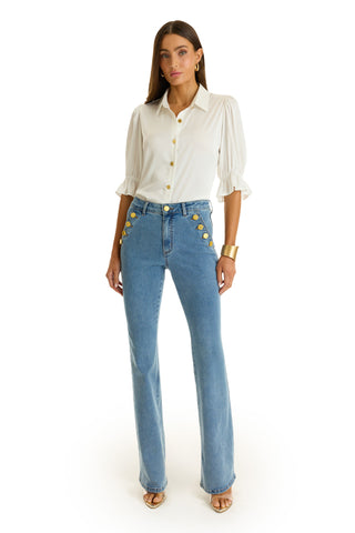 A woman stands facing forward wearing the Cindy Ruffle Sleeve Blouse in white with puffed sleeves, paired with blue high-waisted jeans featuring gold buttons. She accessorizes with sandals, a gold bracelet, and gold hoop earrings.