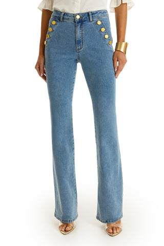 Wearing the Allegra Bootcut Jean in medium blue with gold button front pockets, styled with a white blouse, gold cuff bracelet, and open-toe heels, this look is set against a white background for vintage flair.