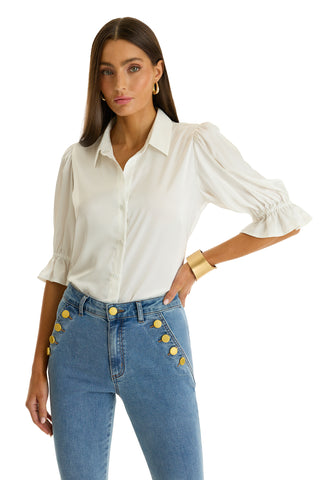 A woman stands facing forward with one hand on her hip, wearing the Cindy Ruffle Sleeve Blouse in white—featuring feminine ruffle sleeves—and blue high-waisted jeans with gold buttons.