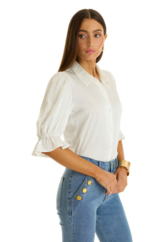 A woman with long brown hair wears the Cindy Ruffle Sleeve Shirt - White, featuring puffed sleeves, paired with blue high-waisted jeans with gold buttons. She stands against a white background, looking left with a neutral expression.
