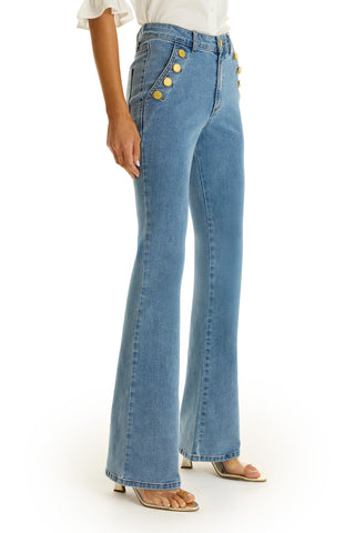 A person wears the Allegra Bootcut Jean in medium blue, featuring gold button front pockets, styled with a white shirt and open-toe heels for a vintage-inspired look.