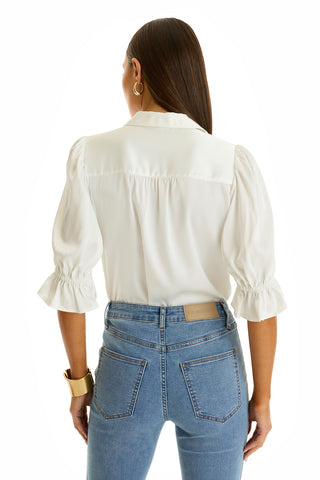 A woman with long brown hair, seen from behind, wears a white Cindy Ruffle Sleeve Shirt and blue high-waisted jeans, styled with gold hoop earrings and a wide gold bracelet.