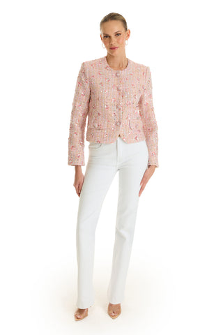 A woman stands against a white background wearing the May Tweed Crystal Jacket in Pink Multi, paired with white straight-leg pants and nude heels. Her hair is pulled back and she stands confidently with one hand on her hip.