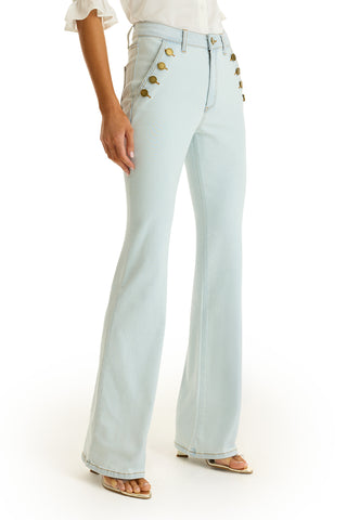 Wearing the Allegra Bootcut Jean - Cloud, light blue with gold-buttoned front pockets, styled with a white blouse and open-toed sandals, a person stands against a white background.