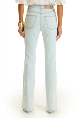 A woman faces away from the camera wearing Allegra Bootcut Jean - Cloud, a white ruffled-sleeve blouse, gold bracelet, and heeled shoes.