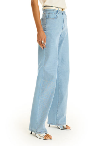 A person wears medium blue Brynn Crystal Jeans, a white belt, white sandals, and a white top. Rings decorate their left hand, which rests at their side. Only the lower torso and legs are shown.