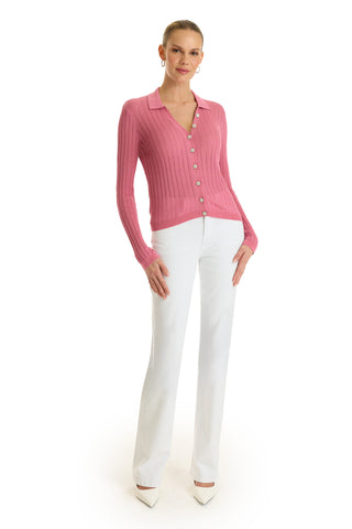 A woman stands against a white background, wearing the Karolina Polo Shirt - Petal Pink with long sleeves and diamond buttons, paired with white pants and pointed-toe heels. Her hair is pulled back and she smiles gently.