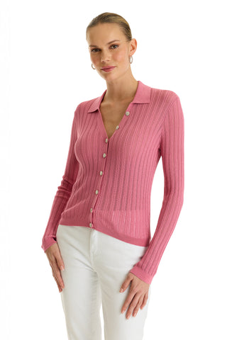 A woman wears the Karolina Polo Shirt in Petal Pink—a lightweight, ribbed, button-up cardigan with diamond buttons and a collar—paired with white pants against a plain white background.