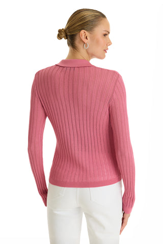 A woman with blonde hair in a low bun wears the Karolina Polo Shirt - Petal Pink with long sleeves and white pants, standing against a white background and looking over her shoulder.