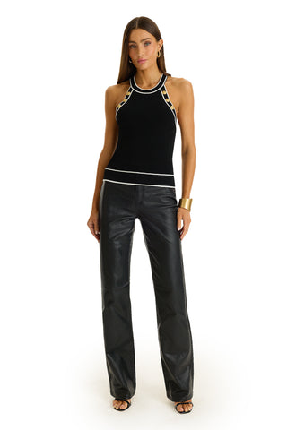 A woman with straight brown hair and a neutral expression stands against a white background wearing the Martha Button Halter Tank - Black, paired with black leather pants, black heeled sandals, and a gold cuff bracelet.
