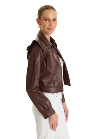 A woman stands sideways, smiling with her hair pulled back, wearing the Chantal Ruffle Sleeve Leather Jacket in Espresso over a light top and white pants. The chic faux leather jacket adds a stylish statement to her fall look against a white background.