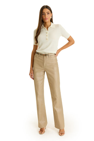 A woman with long brown hair and a neutral expression stands against a white background wearing the Leya Rose Polo Shirt - Pearl, high-waisted tan pants, and open-toe heels, with one hand on her hip.