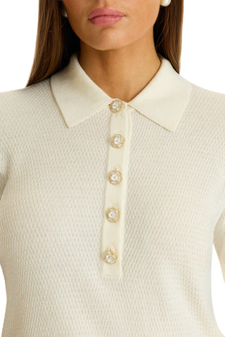 A woman wears the Leya Rose Polo Shirt - Pearl in a soft cream knit with a polo collar and five gold and pearl buttons. Only her lower face and upper torso are shown.