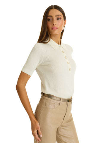 A woman wears the Leya Rose Polo Shirt - Pearl with large buttons and beige high-waisted pants, standing against a white background and looking to her left.