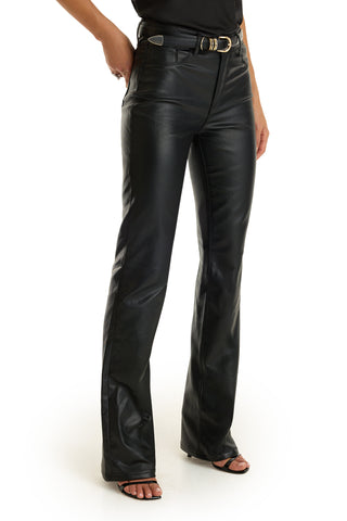 A person wearing the Aria Coated Flare Jean in black, styled with a tucked-in top and heeled sandals, stands against a white background. The jean's modern flare silhouette adds a sleek edge to the ensemble.