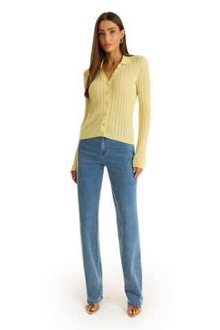 A woman with long brown hair stands facing forward in the Karolina Polo Shirt - Sunshine Yellow, light blue straight-leg jeans, and open-toe sandals, against a plain white background. She has a neutral expression.