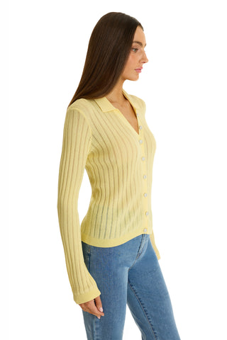 A woman with long brown hair stands in profile against a white background, wearing the Karolina Polo Shirt in Sunshine Yellow and blue jeans—a perfect lightweight knit for spring style.
