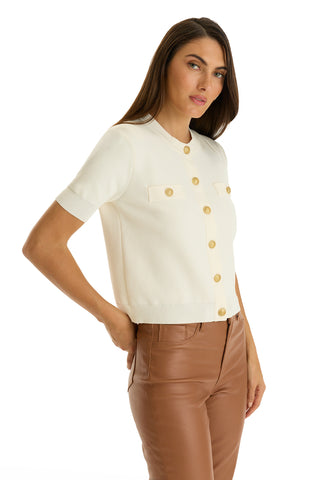 A woman with long brown hair wears the Bonnie Knit Cardigan Top in Pearl, featuring short sleeves, front pockets, and tan buttons, paired with high-waisted brown leather pants. She poses against a white background.