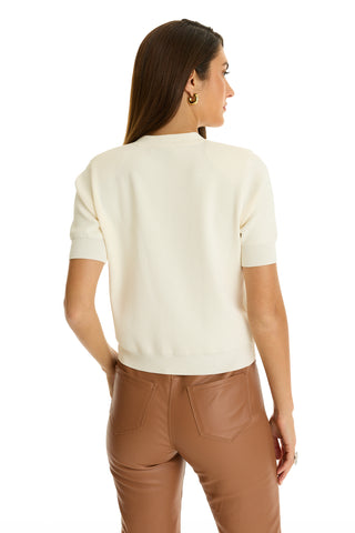 A woman with long brown hair stands facing away, showcasing the back of the Bonnie Knit Cardigan Top - Pearl with gold buttons, paired with high-waisted brown leather pants against a white background.