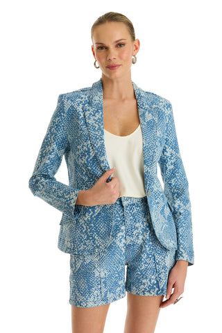 A woman smiles and poses against a white background wearing the Marie Single Breasted Blazer - Crystal Snake, featuring a blue and white snake print with matching shorts and a white top underneath.