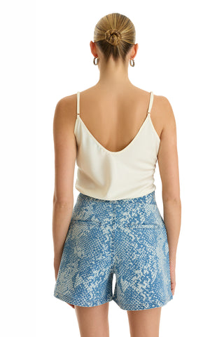 A woman with her hair in a neat bun and hoop earrings faces away, wearing a white sleeveless low-back top and the Beverly Pleated Short - Crystal Snake. The background is plain white.