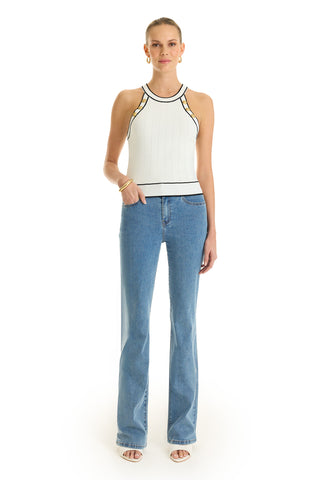 A woman stands against a white background wearing the Martha Button Halter Tank in white with black trim, light blue flared jeans, white sandals, gold hoop earrings and bangles, her hair pulled back.